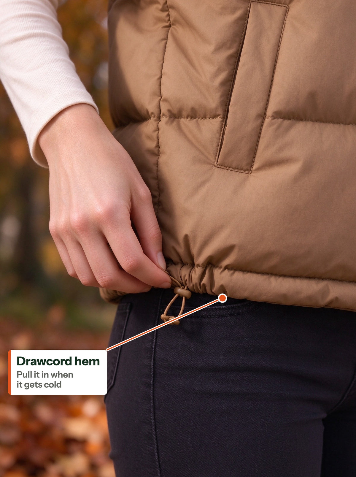 Hand pulling the drawcord toggle at the hem of the camel quilted vest (EN label)