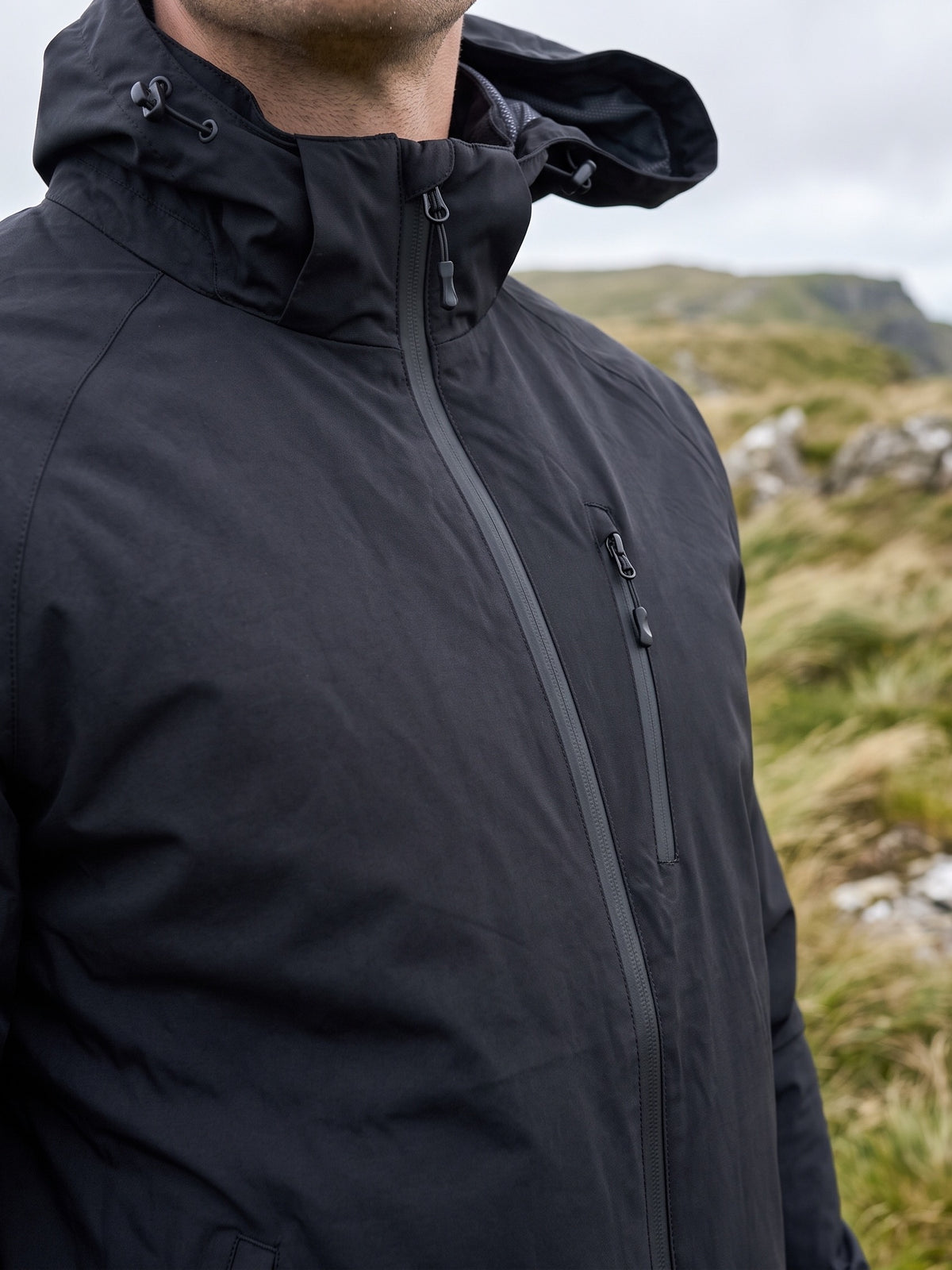 Black wind shell worn on an exposed grassy ridge, the thin polyester face and the attached hood with drawcord at the collar