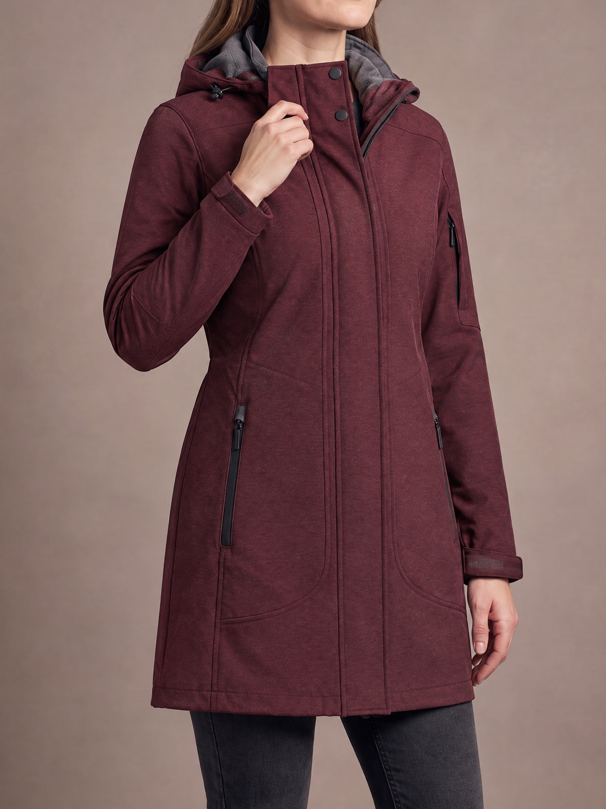 Long fleece-lined softshell coat in burgundy worn with one hand at the snap collar, grey fleece-lined hood and zipped hand pockets