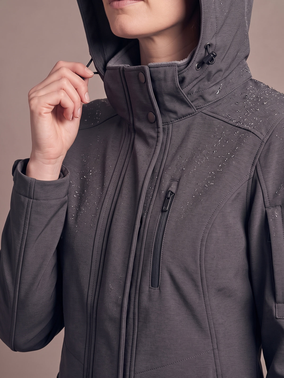 Charcoal softshell coat worn with the grey fleece-lined hood up, hand pulling the hood drawcord, snap stand collar closed and sleet beading on the shoulders