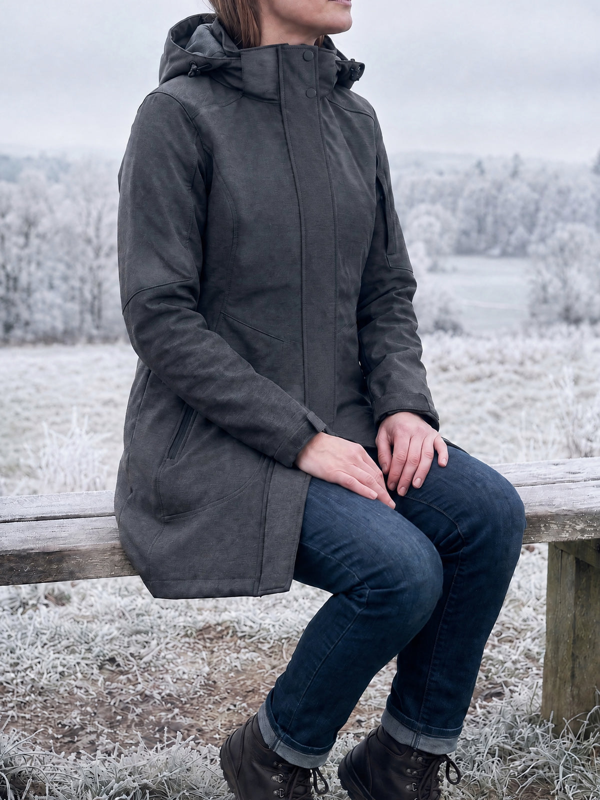 Woman seated on a wooden bench at a frosty viewpoint in the charcoal long softshell coat, zipped and snapped closed, the coat covering hips and thighs