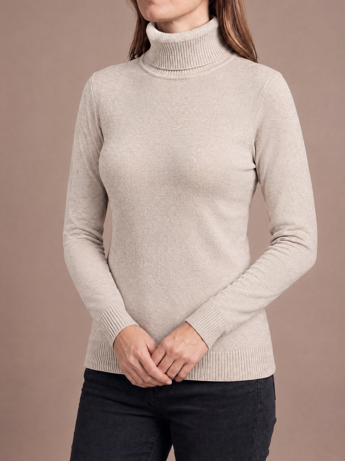 Woman wearing the plush-lined knit turtleneck in oatmeal with hands clasped low in front, fold-over collar, slim fit and ribbed cuffs and hem