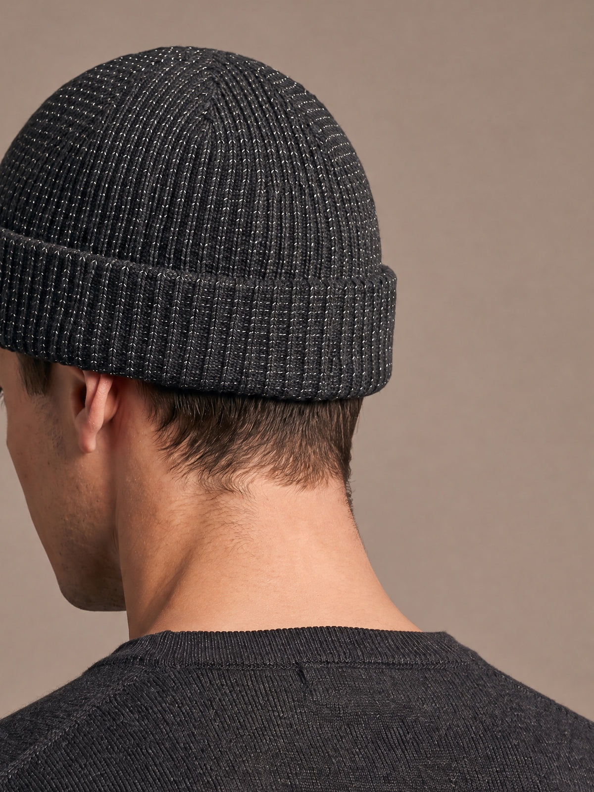 Reflective rib-knit beanie in grey worn by a man seen from the side-back with the head tilted slightly down, short crown, folded cuff and reflective flecks