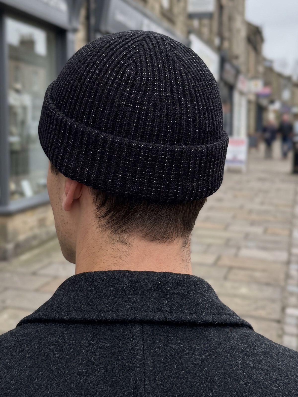 Man seen from behind on a town high street in grey daylight wearing the black rib-knit beanie with a dark wool coat, the reflective yarn only a faint fleck in the knit