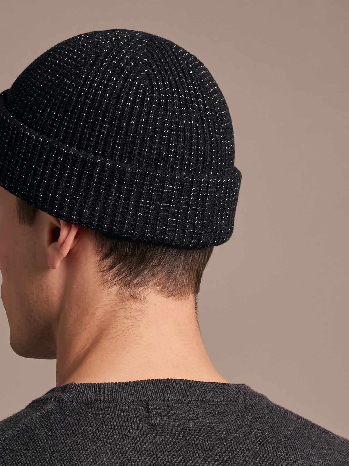 Black reflective rib-knit beanie worn by a man, seen from the side-back, showing the short crown, folded cuff above the ear and fine reflective flecks in the knit