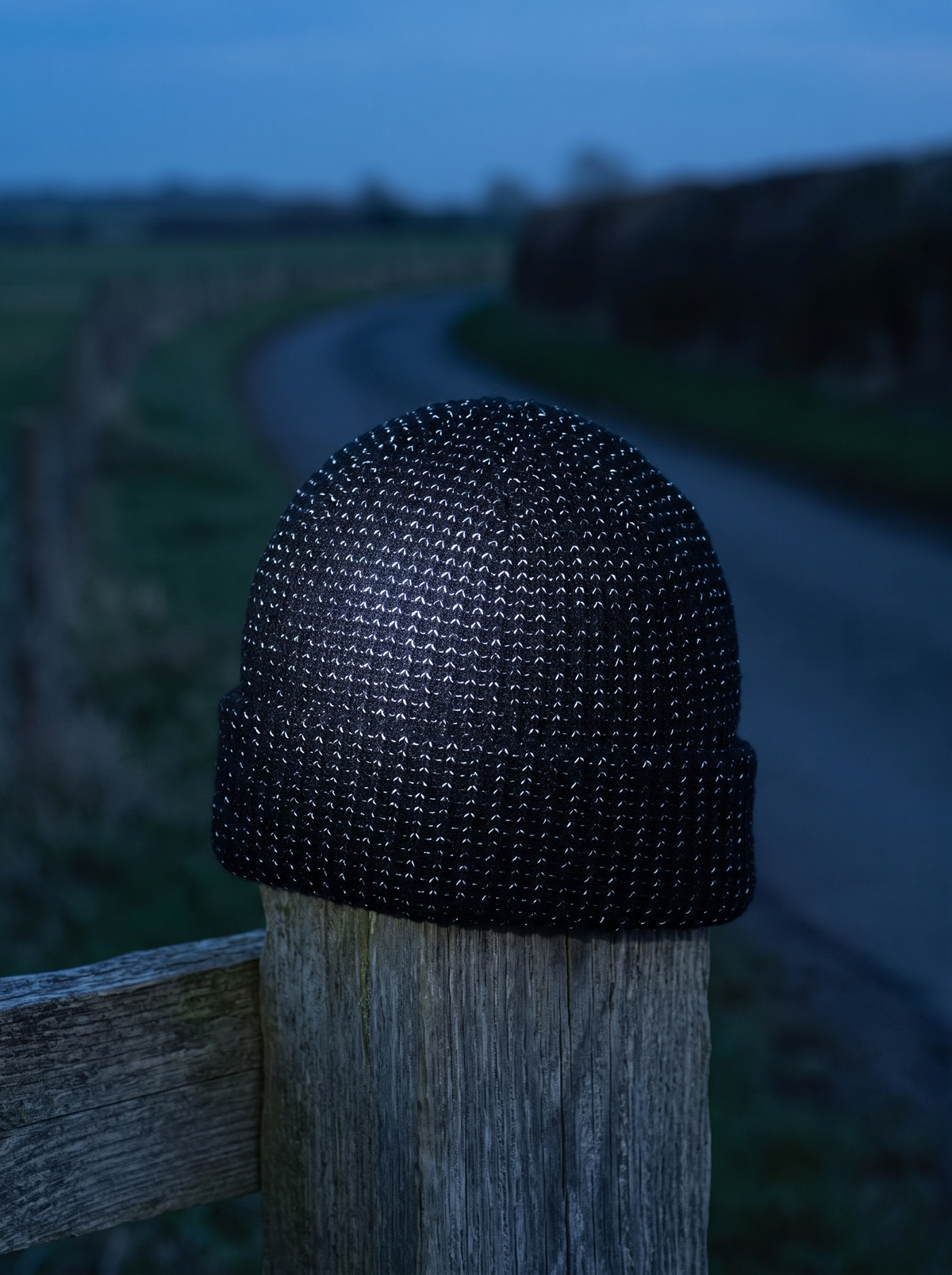Black rib-knit beanie on a fence post at dusk with its reflective yarn lighting up under a torch beam