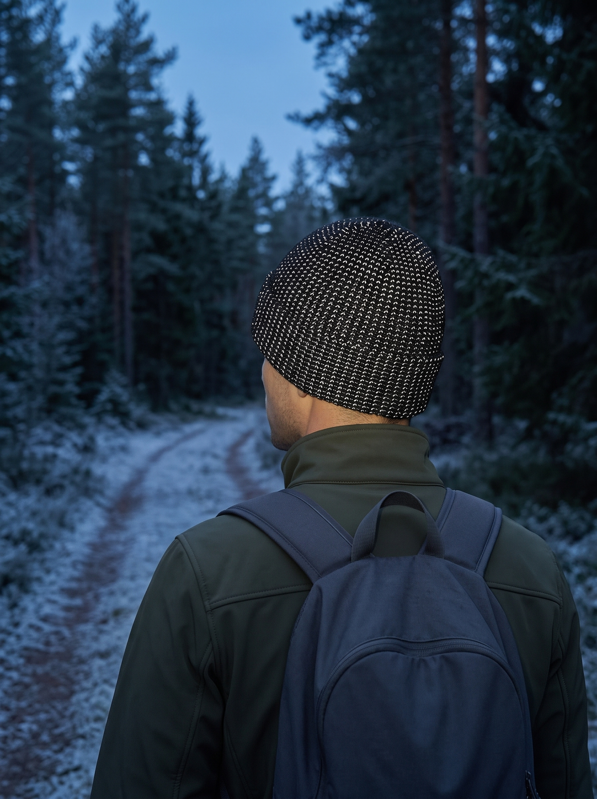 Man seen from behind on a frosty forest trail at dusk, the reflective yarn of the black beanie lighting up silver-white