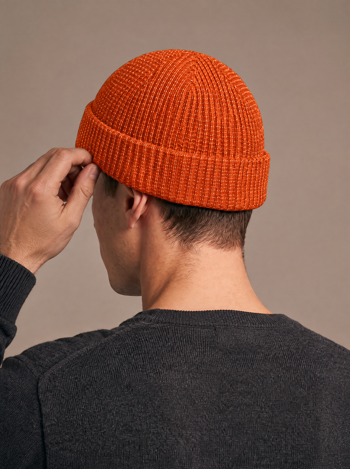 Reflective rib-knit beanie in orange worn by a man seen from the side-back, one hand adjusting the folded cuff, reflective flecks in the knit