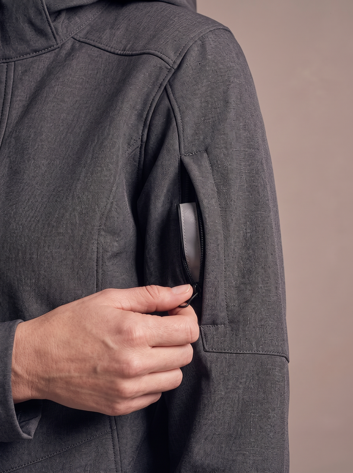 Hand opening the zipped pocket on the upper sleeve of the charcoal softshell coat, a reflective band showing inside