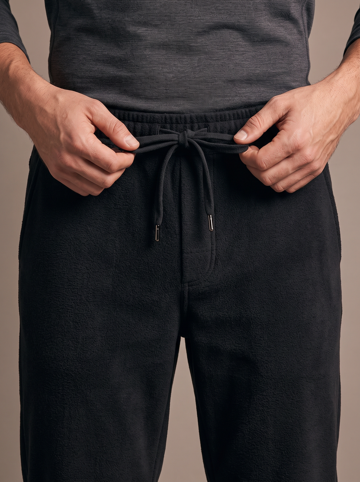 Hands tying the drawcord at the elastic waistband of the black double-face fleece jogger, base layer hem above