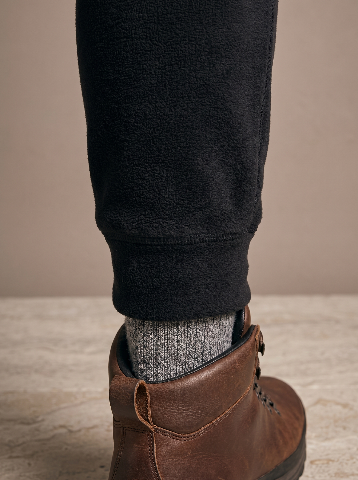 Ribbed ankle cuff of the black fleece jogger closing over a grey wool sock above a brown leather walking boot