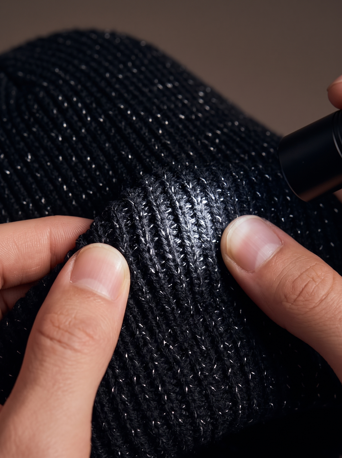 Macro of the black rib knit stretched between two fingers, fine reflective yarn flecks glinting in a torch beam