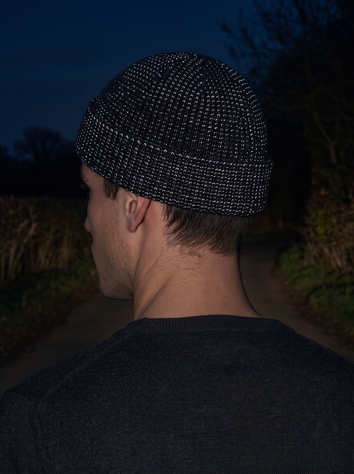 Black rib-knit beanie seen from behind on a dark country lane, the reflective yarn in the knit glinting silver-white in a headlight beam