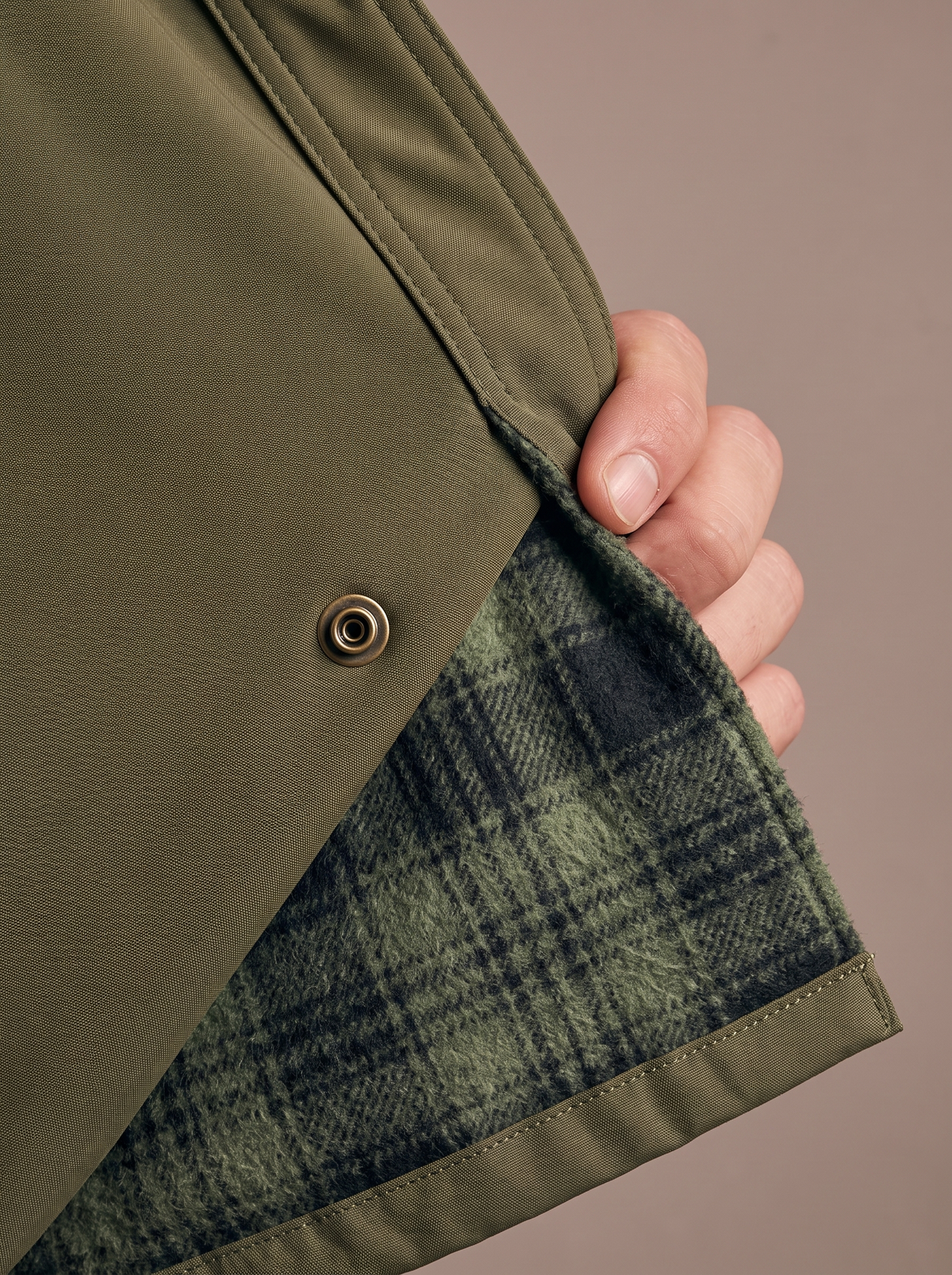 Macro of the front edge folded back: smooth army green polyester outer with a brass snap over the soft brushed green plaid flannel lining