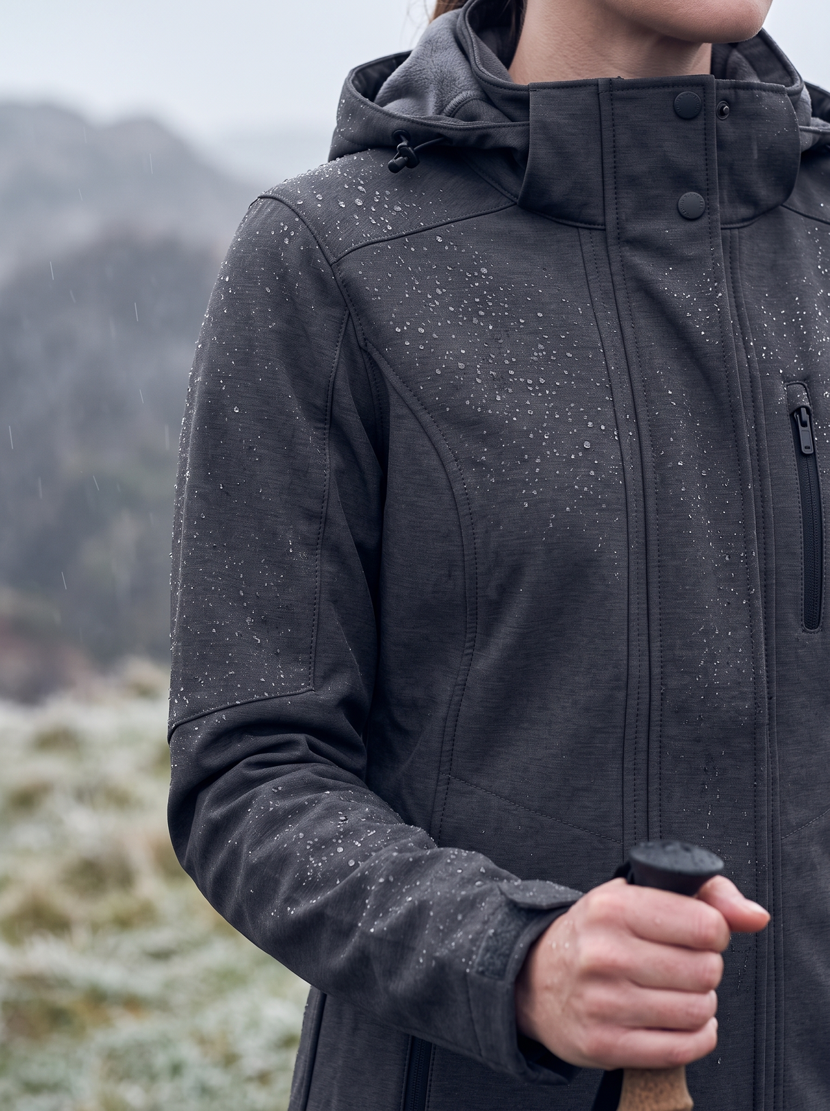 Drizzle beading on the charcoal heather stretch softshell at the shoulder and chest, worn at a frosty viewpoint with the grey fleece-lined hood down