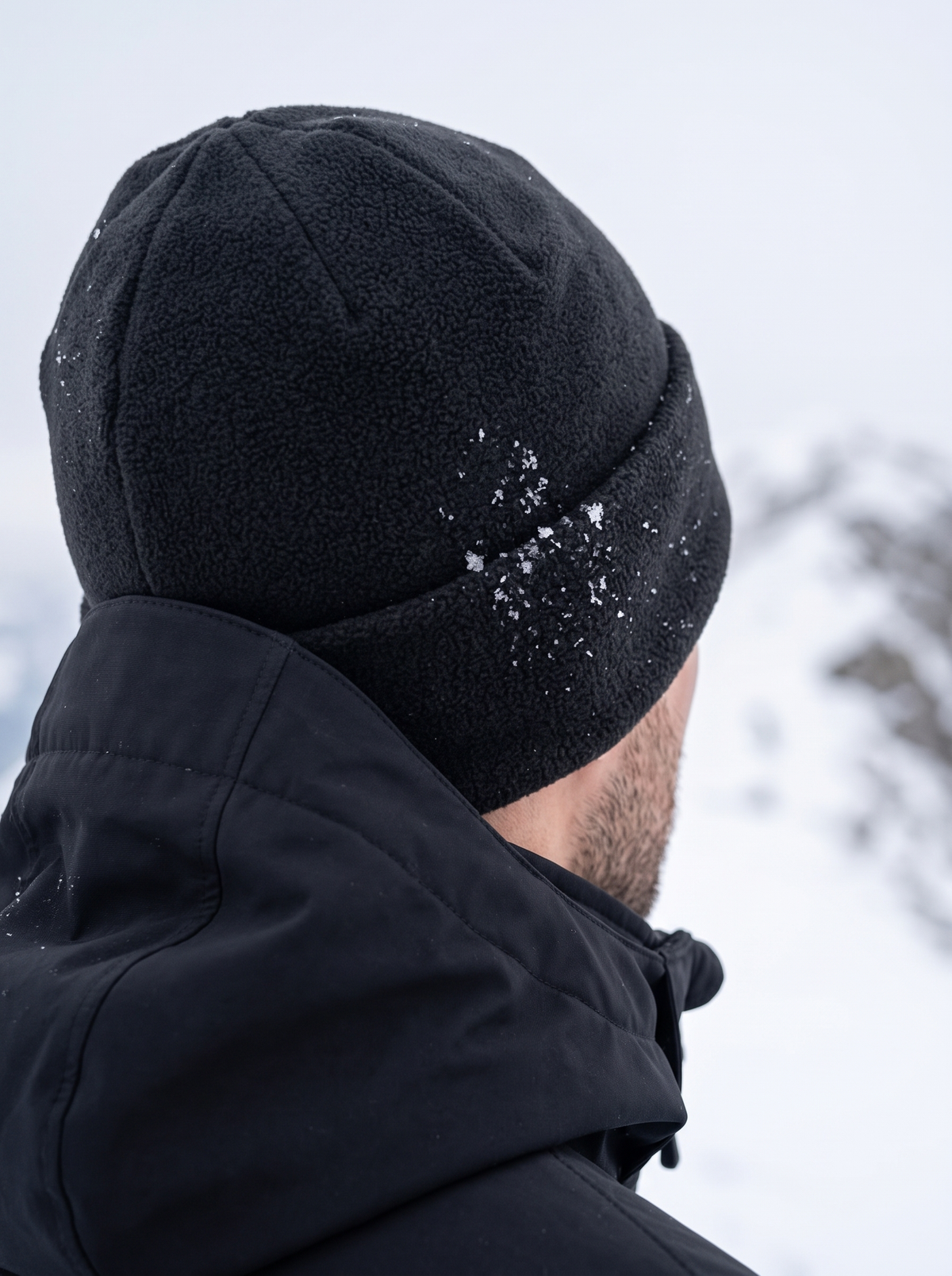 Black polar fleece beanie with a folded cuff seen from behind on a snowy ridge, snowflakes caught in the fleece