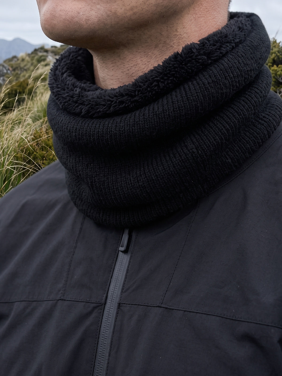 Black knit neck gaiter worn on an exposed ridge, the fleece lining folded over at the top edge against the jaw