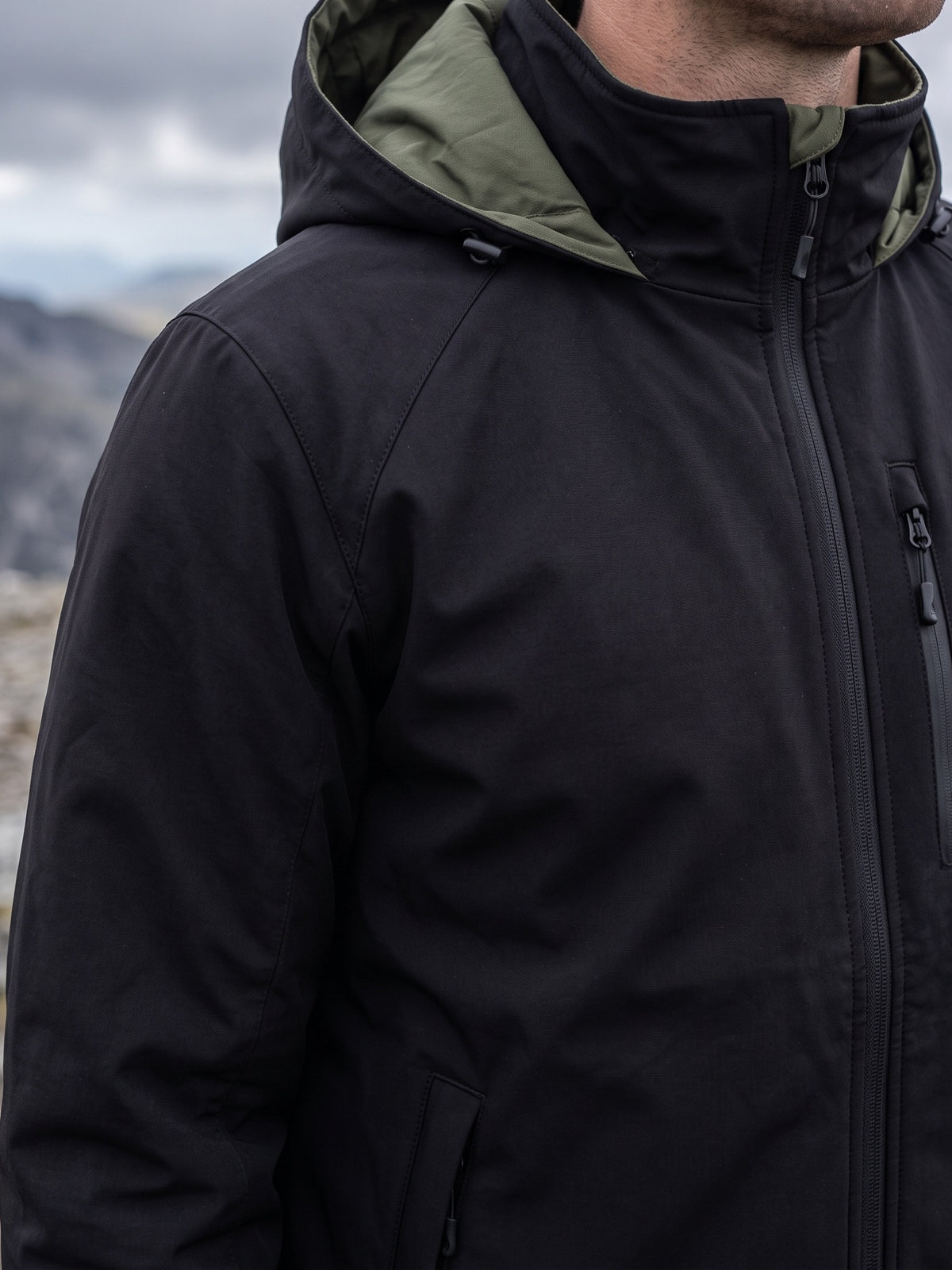 Black hooded softshell worn on a windy rocky ridge, the matte stretch face at the shoulder and the olive hood lining open at the collar